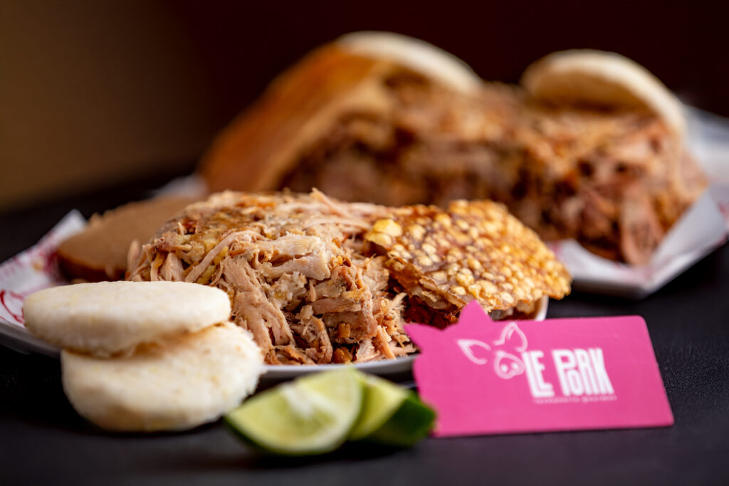 Porción de lechona servida con arepas y limón sobre plato gourmet, acompañada de tarjeta de presentación Le Pork.
